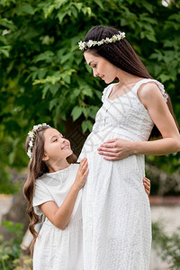 快乐的怀孕母亲和可爱的微笑女儿穿着白色连衣裙和花圈站在一起图片