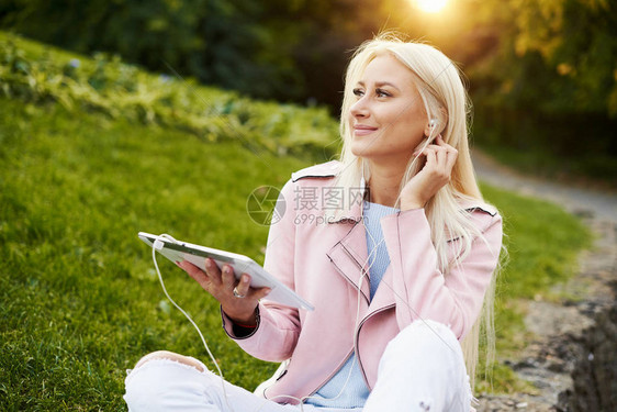 微笑的英俊女人在公园里用她的手机或平板电脑在耳机里听轻松的音乐一名学生听在线讲座并准备考试享图片