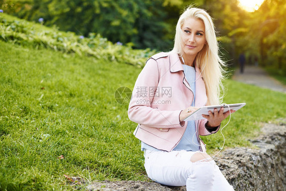 微笑的英俊女人在公园里用她的手机或平板电脑在耳机里听轻松的音乐一名学生听在线讲座并准备考试享图片