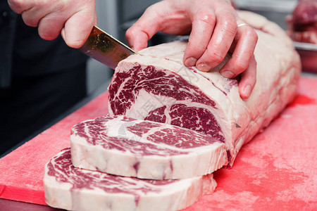 特写宏厨师的手切ribeye大理石牛排用刀在餐厅厨房金属桌子上的红色塑料切板上概念包装半成品背景图片