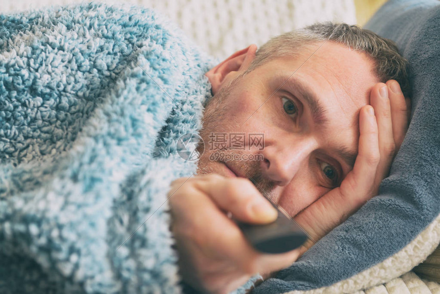 患有抑郁症的成熟男子在床上和看电视时被毯子覆盖着图片