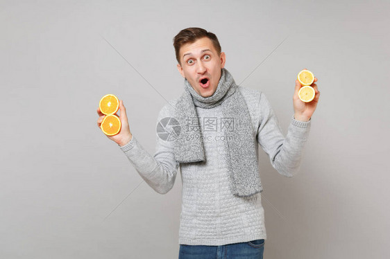 穿着灰色毛衣围巾张开嘴巴拿着灰色背景中突显的橙子柠檬的年轻人感到惊讶图片
