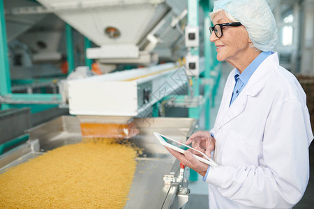 在工厂作的微笑高级女站在传送带旁并使用数字平板电脑控制食品生产复制图片