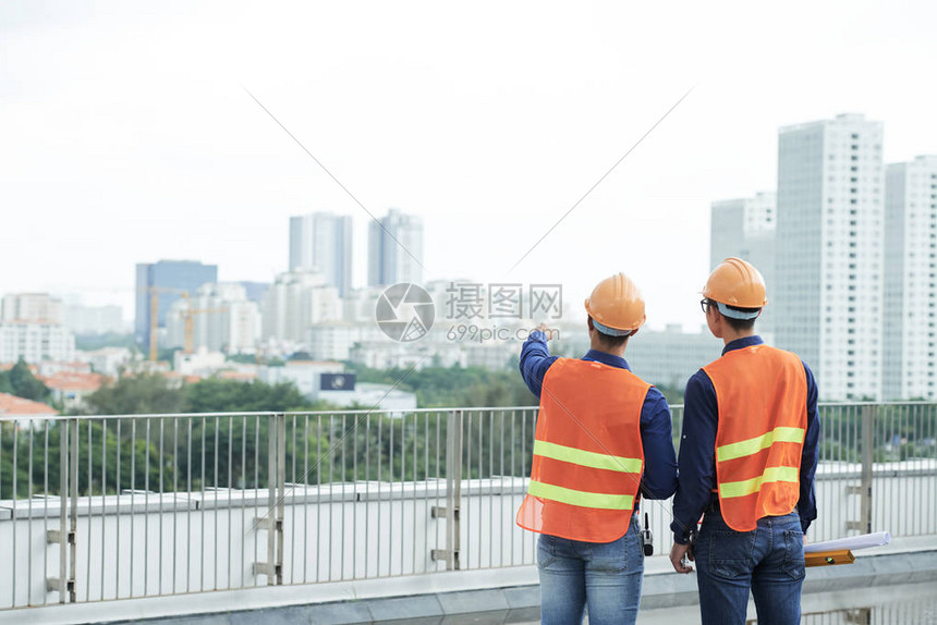 身戴头盔和腰衣的两个人背视镜站在建筑工地上图片