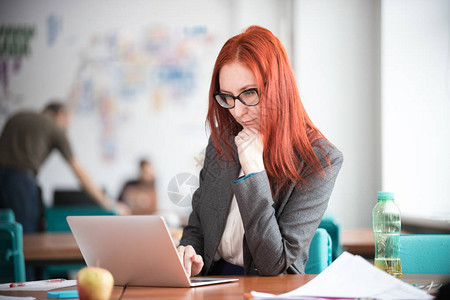 坐在办公室和笔记本电脑上工作的身着眼镜的生姜体贴的女商图片