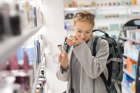 金发碧眼的年轻女旅行者穿着外套和旅行背包在机场免税店选择香水休闲女士在美容店随时地测试图片