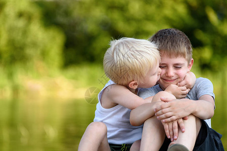 两个小兄弟坐在户外一个亲吻另一个的脸颊远处的绿树模糊友谊图片