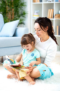 漂亮的年轻母亲正在给坐在房间地板上的地毯上的女儿看书和图片