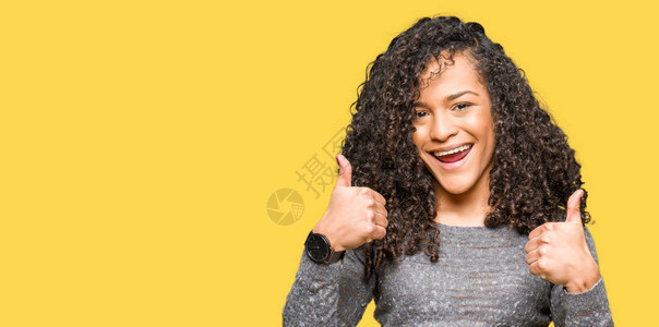 穿着灰色毛衣的卷发年轻美女成功标志用手大拇指微笑和快乐地做积极动作图片
