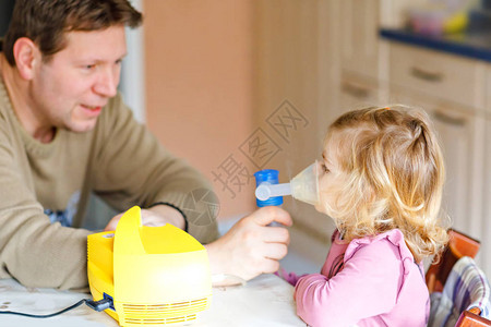 蹒跚学步的小女孩在家里用雾化器吸入父亲帮助和拿着设备孩子有流感咳嗽和支气管炎哮喘吸入器吸背景图片