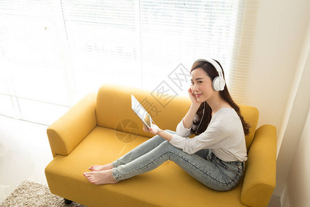 美丽的亚洲年轻女用耳机在房间的沙发上用平板电脑听音乐图片