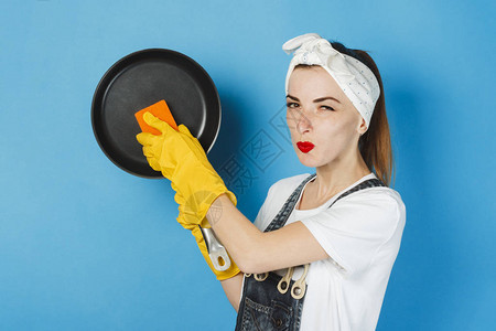一个面带微笑手帕和黄色橡胶手套的年轻女孩在蓝色背景上擦拭煎锅清洁理图片