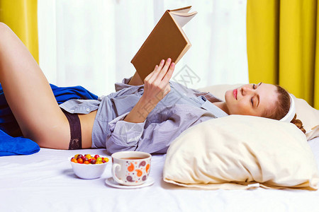 感的金发女孩在床上放松耳机列表音乐和阅读书籍糖豆和她附近的热茶家庭生活朴素背景图片