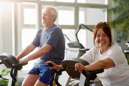 快乐的老年女子在健身房工作在健身房里微笑的老夫妇们骑固定自图片