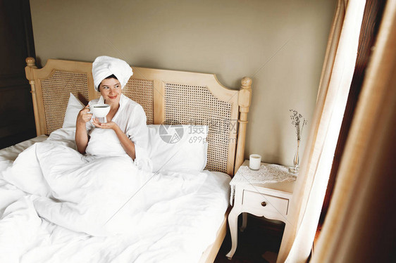 美丽快乐的年轻女子在酒店房间或家庭卧室的床上喝咖啡时尚的黑发女孩穿着白毛巾享受早晨的热图片