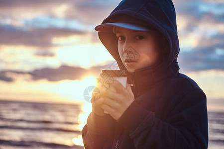 穿着帽子和兜帽的漂亮小男孩在海边喝茶图片