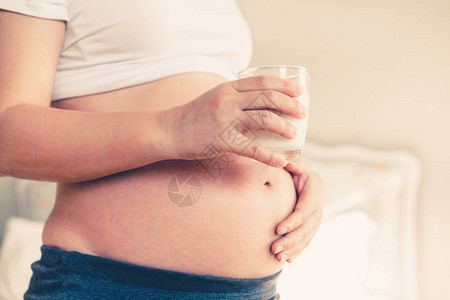 快乐的孕妇在照顾孩子的同时在家喝玻璃杯牛奶年轻的准妈抱着婴儿在怀孕的肚子里钙质食物营养有图片