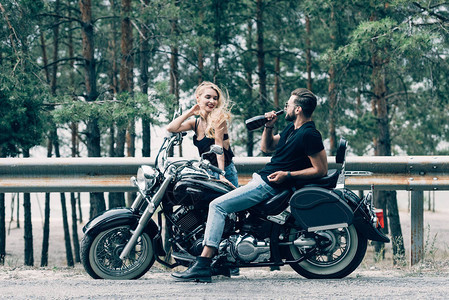 坐在黑色摩托车上和男人在绿林附近路上喝瓶酒喝的年轻骑自行背景图片