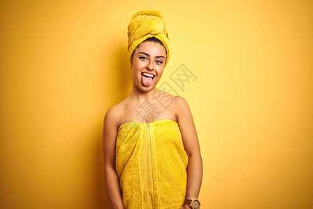年轻美女洗完澡后穿着毛巾在孤立的黄色背景下带着可笑的表情用舌头说出快乐的话图片