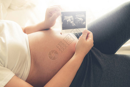 孕妇在照顾孩子的同时在家感到快乐年轻的准妈拿着照片产前护理和图片