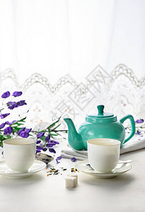 两个带绿茶的白茶杯茶色壶白色背景上的紫色花前视图片