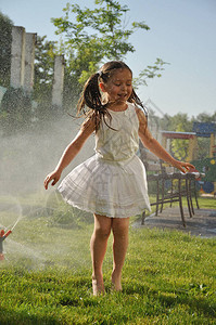 孩子们在炎热的夏天玩水女孩在绿色草坪上的喷水下奔跑带花园洒水器的孩图片
