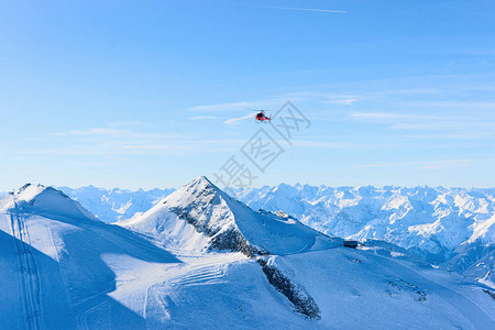 冬季在阿尔卑斯山的奥地利蒂罗尔州齐勒河谷的辛特图斯冰川滑雪胜地上空飞行红色直升机斩波器和高山与雪飞机和蓝天图片