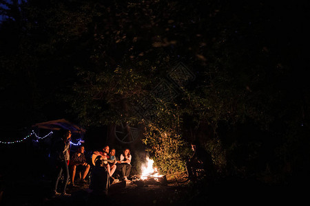 时髦的男人在里的夜间营地的大篝火上演奏原声吉他图片