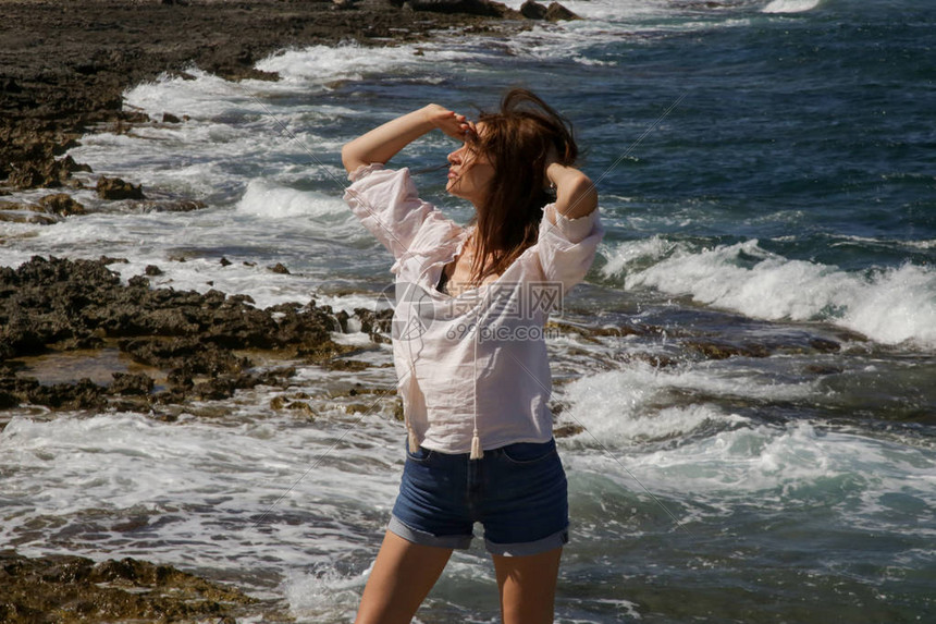 身穿白衬衣和牛仔短裤的年轻美女在风寒雨中的岩石海岸享受暑假在远离旅游景点的偏远地图片