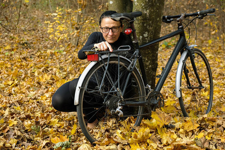 一名身着黑衣和眼镜的58岁妇女坐在一辆自行车附近的黄色叶子表面图片