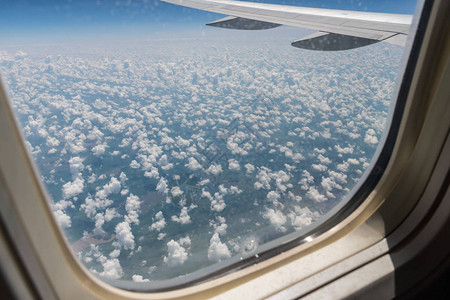 这是从飞机翼上的飞机舷窗下面的地面和飞行中夏日的云层看到的景色图片