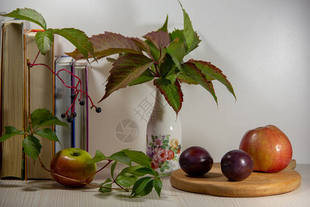 秋天的静物与书一个花瓶五颜六色的叶子木板上的苹果和李子白色背景上图片