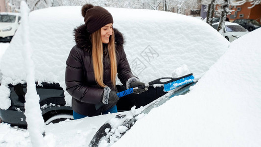 穿着棕色大衣和帽子微笑的漂亮女孩在下雪后用刷子擦洗汽图片