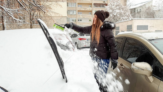 穿着夹克的微笑女孩的镜头用刷子清理积雪覆盖的汽车在初雪的冬日早晨刮擦挡图片