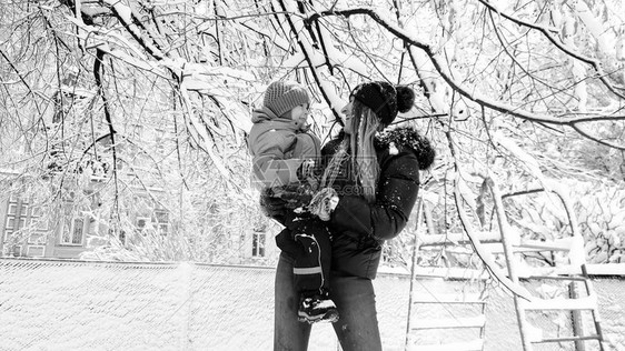 在公园操场玩弄雪花覆盖的树上玩耍穿着夹克和帽子带着可爱儿子图片