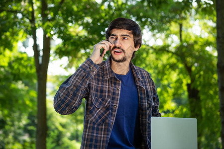 长着胡子和胡子的棕发青年自由职业者在公园里坐在笔记本电脑上讲智能手机图片