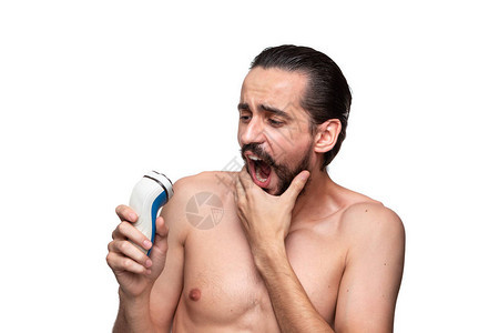 吓坏了留着胡子的大胡子男人看着电动剃须刀光秃地站在白色背景上早晨治疗和剃须的概念是时候修剪你的胡须了早图片