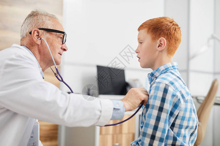 高级医生在儿童保健诊所咨询时用听诊器听可爱红发男孩心跳和呼吸的侧