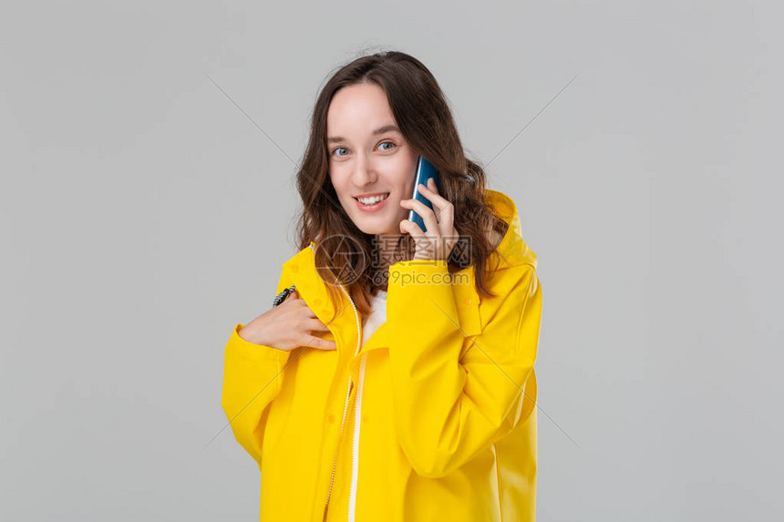 穿着黄色雨衣的漂亮的黑发女人看着快乐地在智能手机上聊天隔着灰色背景被孤立了图片