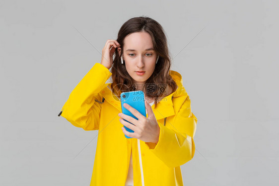 穿黄色雨衣的漂亮的黑发女人正在用智能手机做自拍隔着灰色背景模型戴无线耳膜移动摄影图片