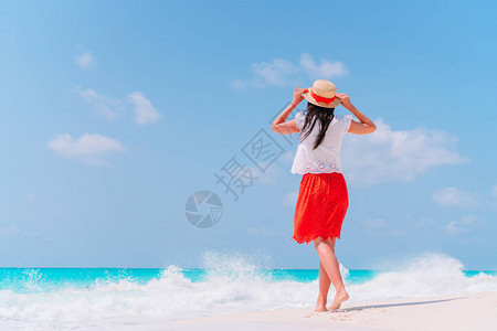 穿着红裙子和帽子的快乐女孩在白色沙热带海滩上行走图片