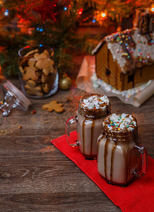 两杯可加棉花糖生奶油和巧克力糖浆放在木桌和姜饼屋饼干罐和圣诞树上图片