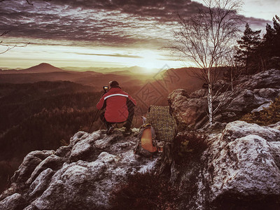 红色夹克摄影师和编织帽的摄影师在悬崖上的三脚架和思维上图片