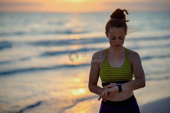 日落时在海边穿着风格衣服的年轻女使用智能手表在健身应用程图片