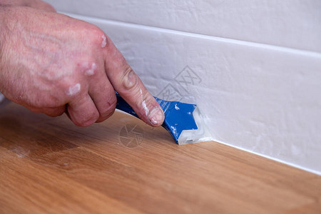 用蓝色刮刀将白色密封剂接缝化合物填缝剂涂在木桌面和米色瓷砖墙的接缝上的专业水背景图片
