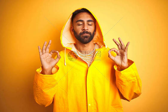 身穿雨衣的印度青年男子带着头罩站在孤立的黄色背景上放松图片