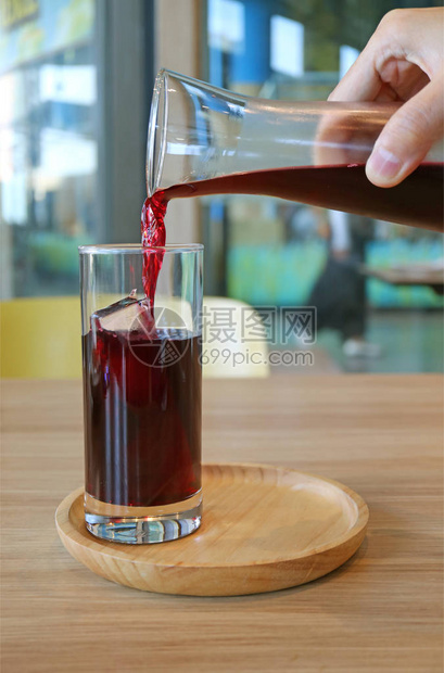 一只手将深红彩色Roselle果汁倒入木质托盘上的冰立方玻图片