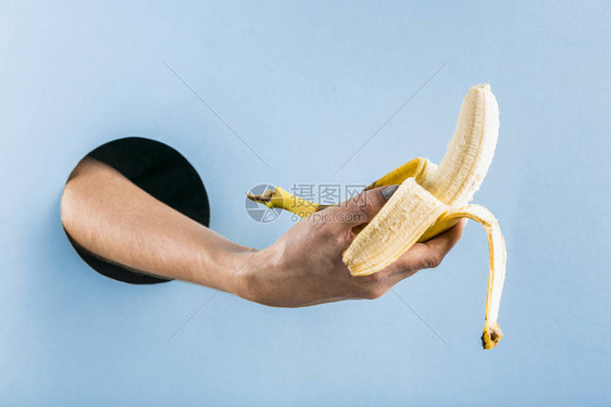 女人的手从蓝纸墙上的黑洞里拉出一块皮香蕉健康的食物图片