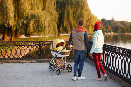 年轻的爱家带着婴儿车在湖边散步微笑的父母在秋天的公园里带着婴儿车爱父母身份家庭季图片