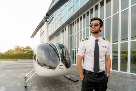 身着白色船长制服的英俊商业飞行员的肖像站在小型私人直升机附近图片